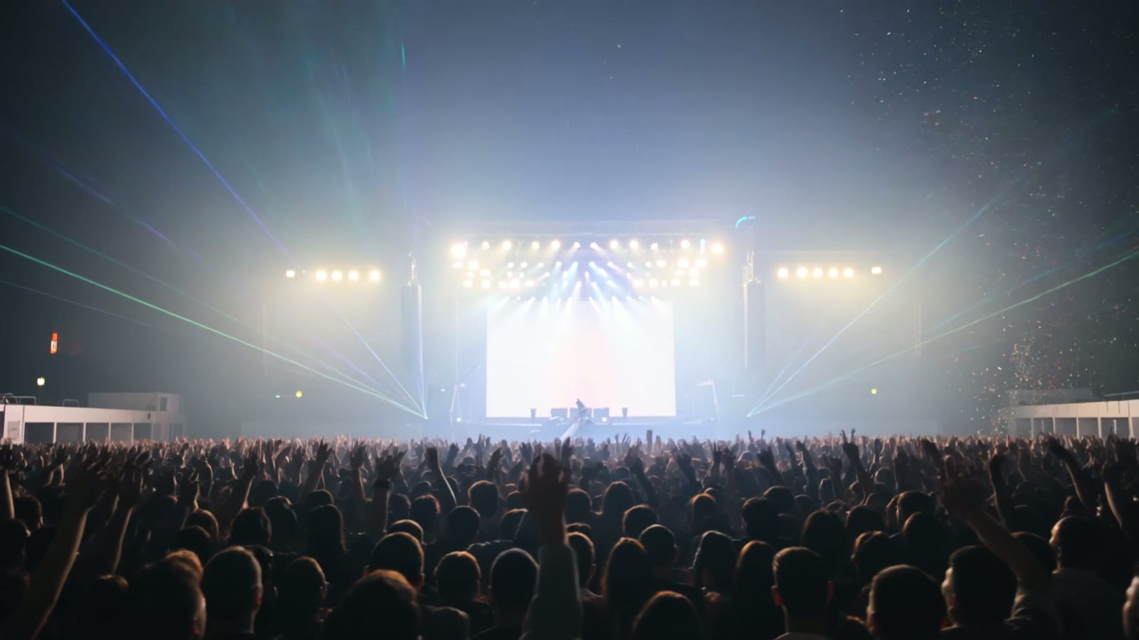 A massive concert crowd with hands raised, silhouetted against a sweeping stage light show, a still from an AI-generated concert clip.