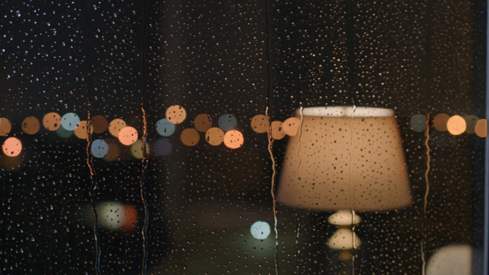 Heavy rain streaming down a window at night with warm blurred city lights glowing beyond the glass, a still from an AI-generated cozy rain clip.